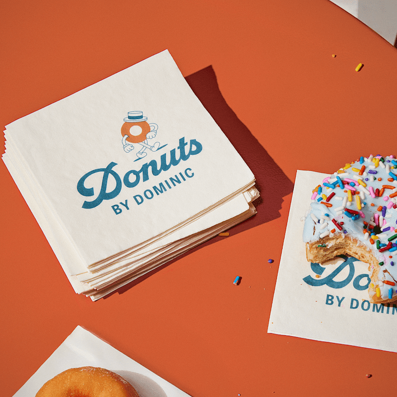 Voqado Paper napkins: Napkins printed for a donut shop, stacked beside a frosted donut with sprinkles